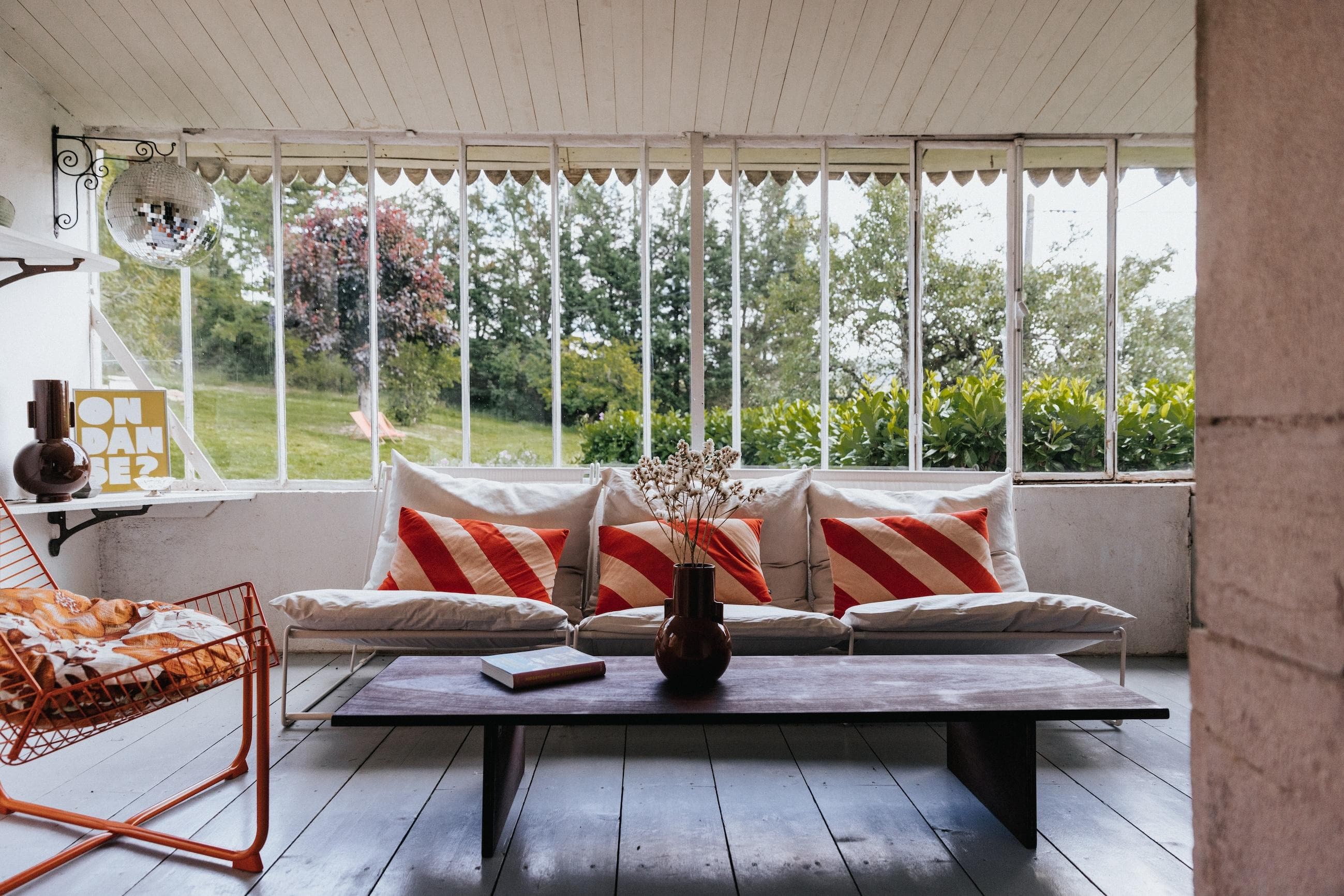 A bright sunroom with large windows, a white sofa with red-striped pillows, a low dark coffee table with vases and a book, and a metal chair with cushions. Trees and greenery are visible outside.