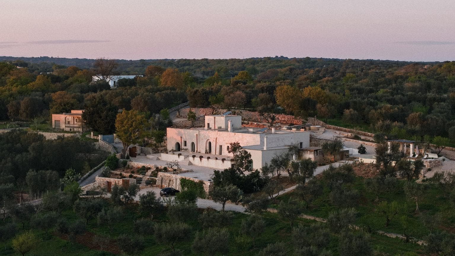 A large, white villa surrounded by trees and greenery sits on a hillside at sunset, with stone terraces, winding paths, and distant forest in the background.