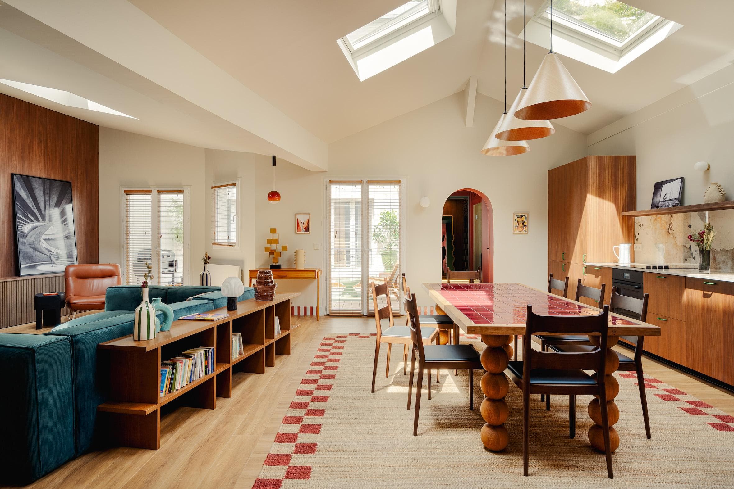 A modern open-plan living and dining area with wooden furniture, a green sofa, skylights, and ample natural light. The space features unique geometric decor, plants, and a kitchen with wood cabinets.