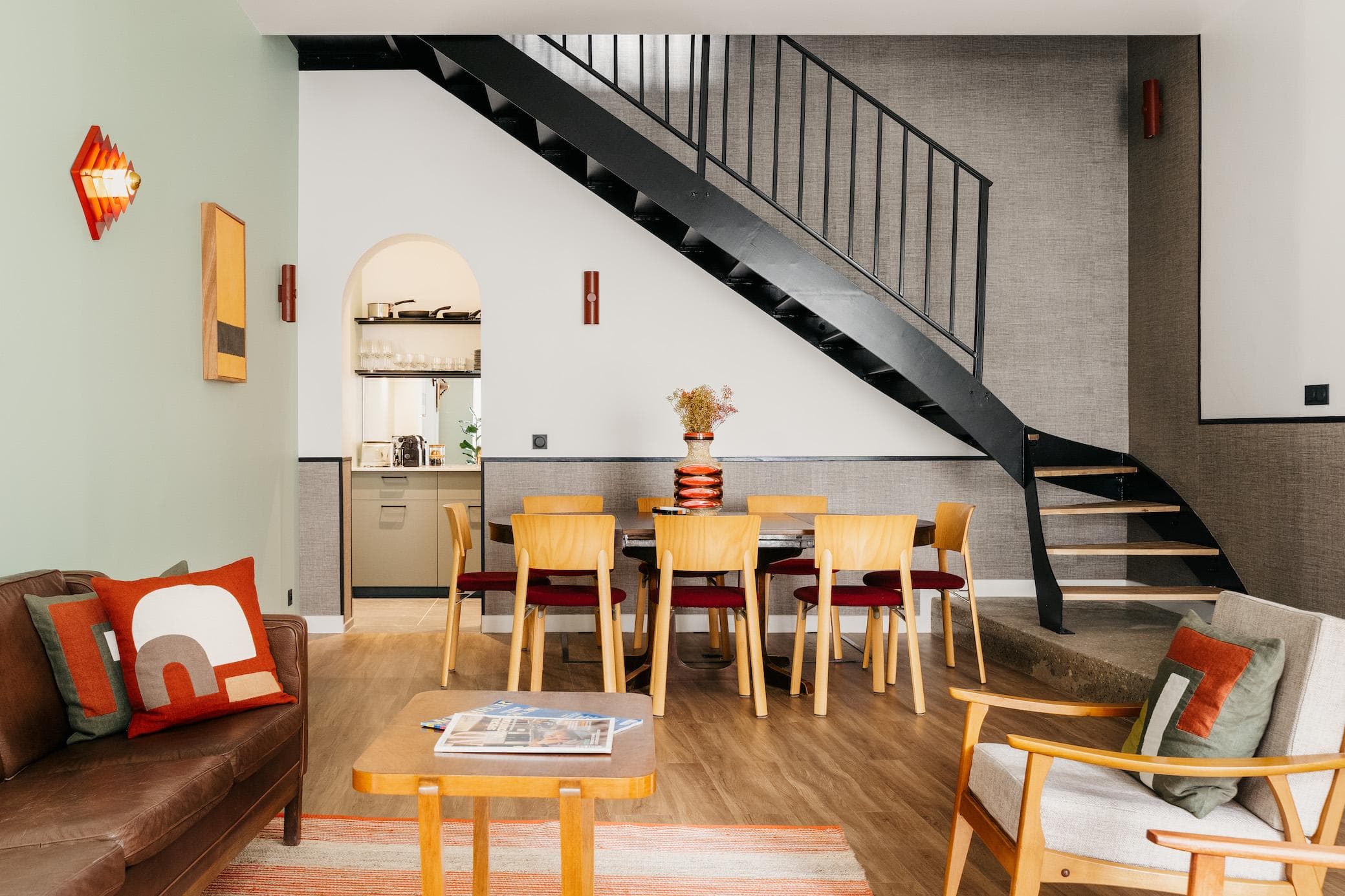 Modern living and dining area with wooden furniture, a brown leather sofa, an armchair, a dining table with chairs, a striped rug, and a black metal staircase against a light and dark gray wall.