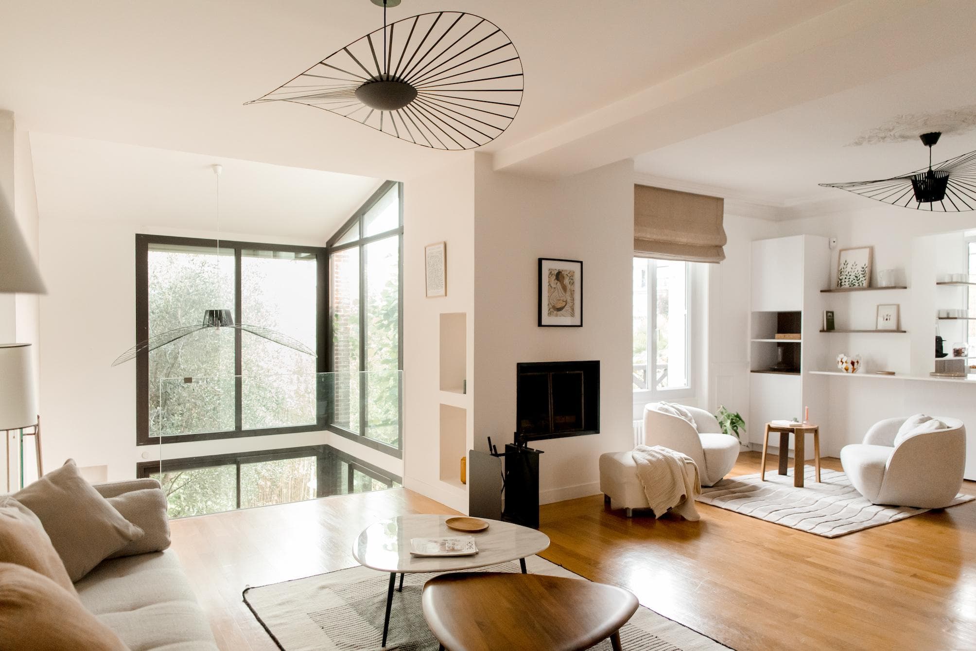 A modern, bright living room with large windows, light wood floors, neutral furniture, a marble coffee table, and black geometric light fixtures. Natural light fills the space, highlighting minimalist decor.