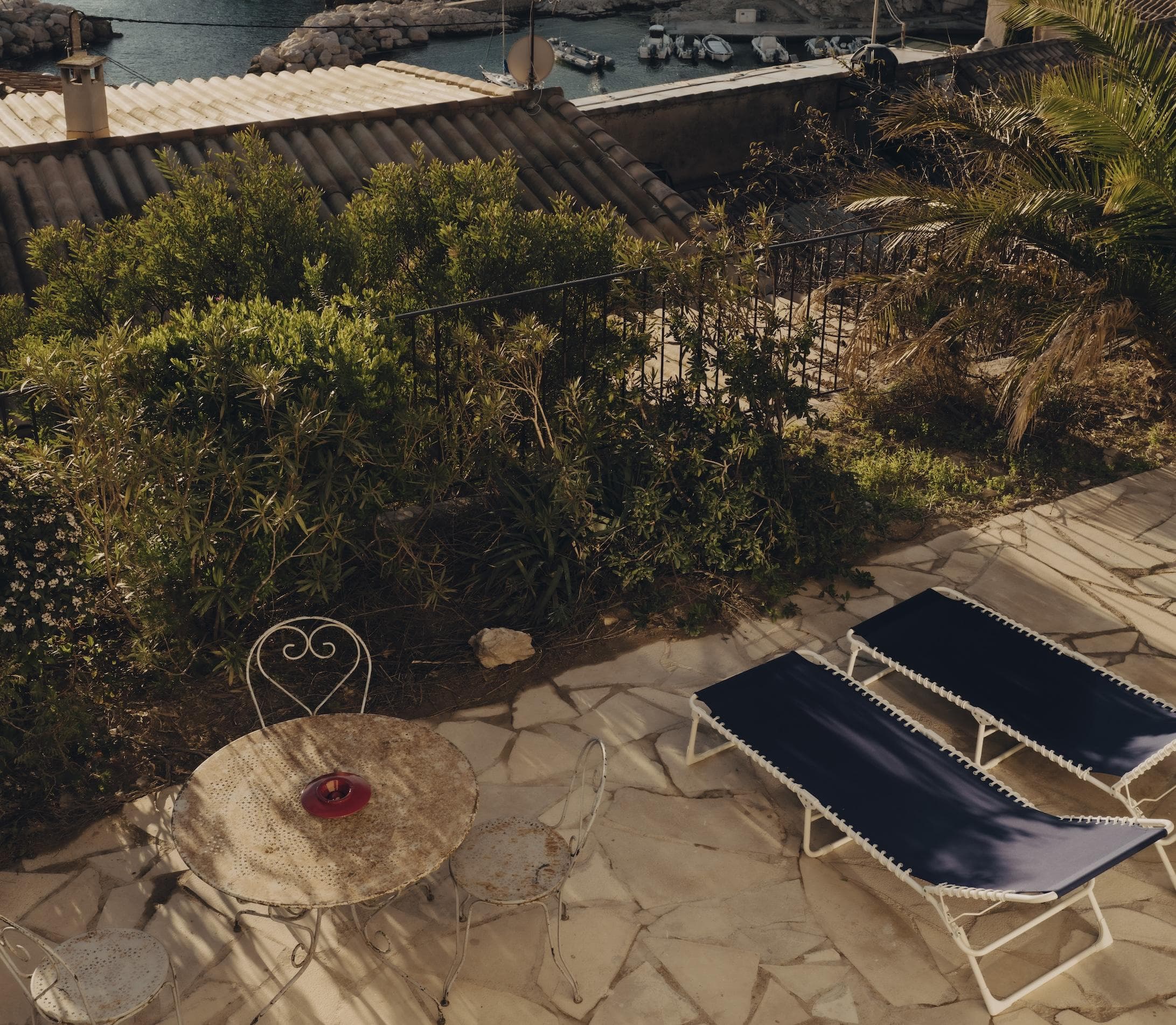 A sunlit stone patio with two blue lounge chairs, a small round table with two chairs, and a bowl containing apples. Lush greenery surrounds the patio, and rooftops and water are visible in the background.