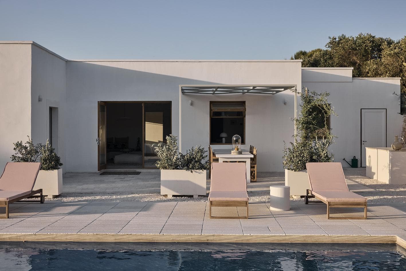 A modern white villa with large windows and a shaded patio, featuring outdoor dining furniture and pink lounge chairs by a swimming pool, set against clear blue skies and greenery.