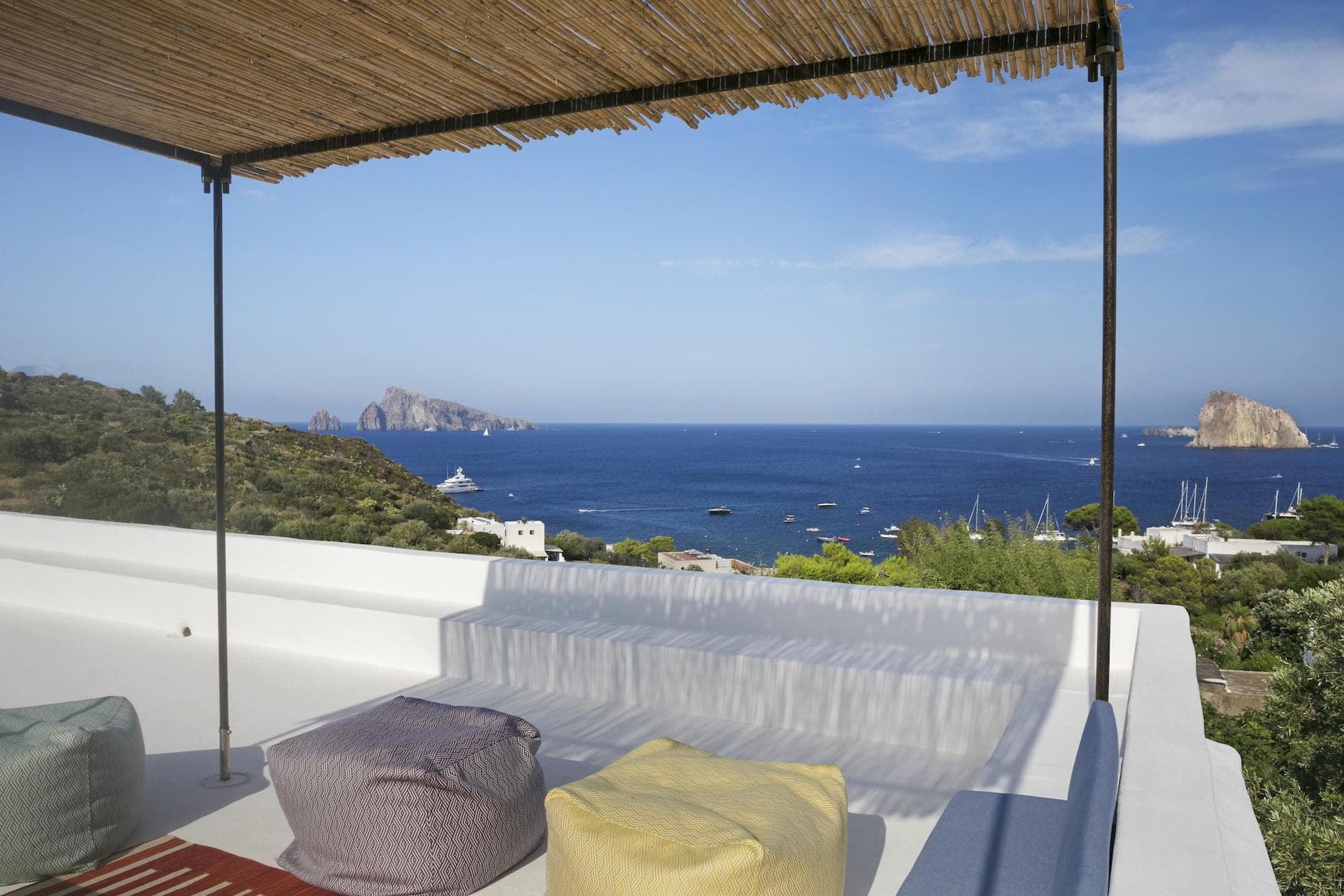 Rooftop terrace with cushioned seating overlooks a blue sea, green hills, boats, and distant islands under a clear sky, shaded by a bamboo pergola.