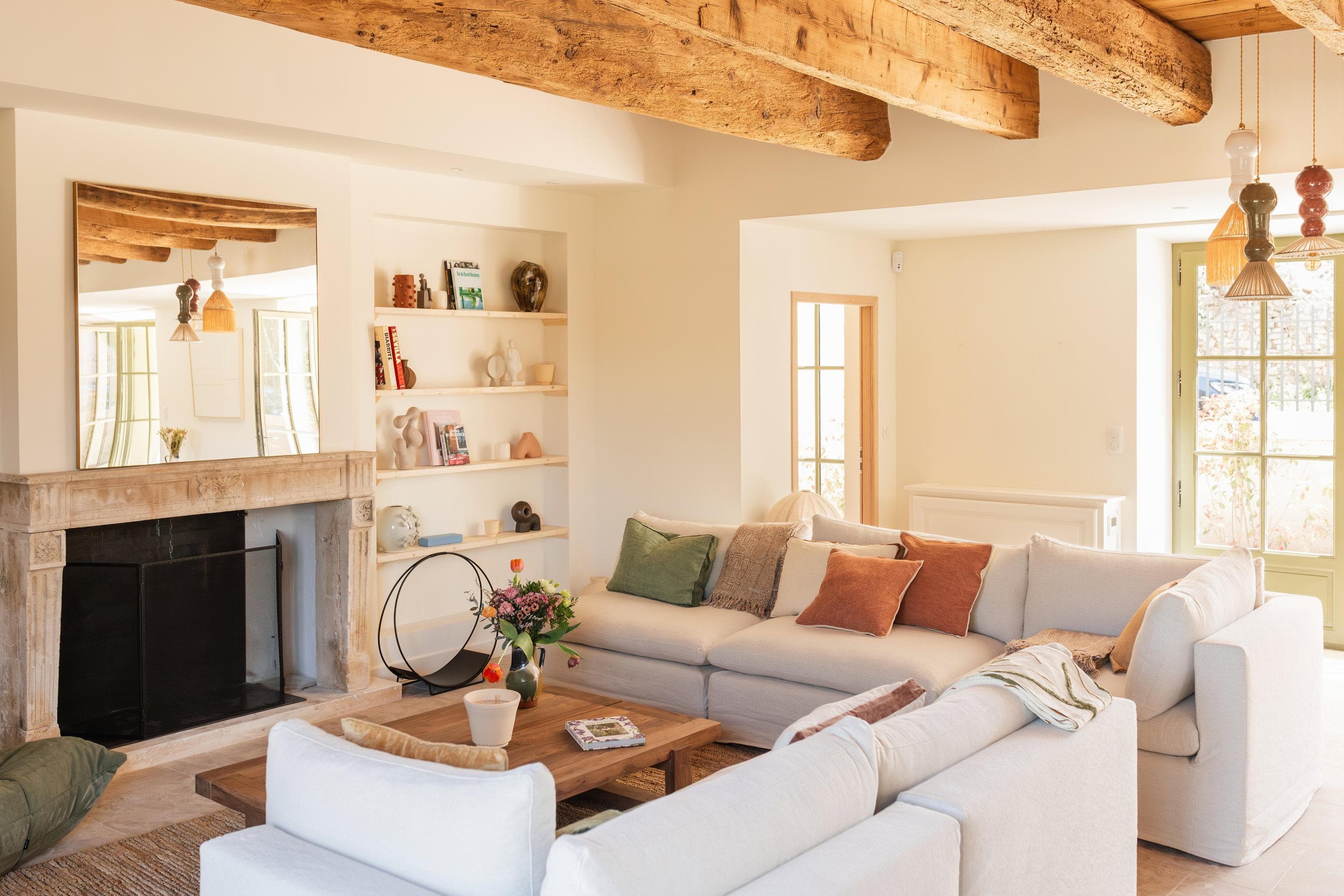 Bright, cozy living room with wooden ceiling beams, a large white sectional sofa, a rustic fireplace, built-in shelves with books and decor, a wooden coffee table, and sunlight streaming through French doors.