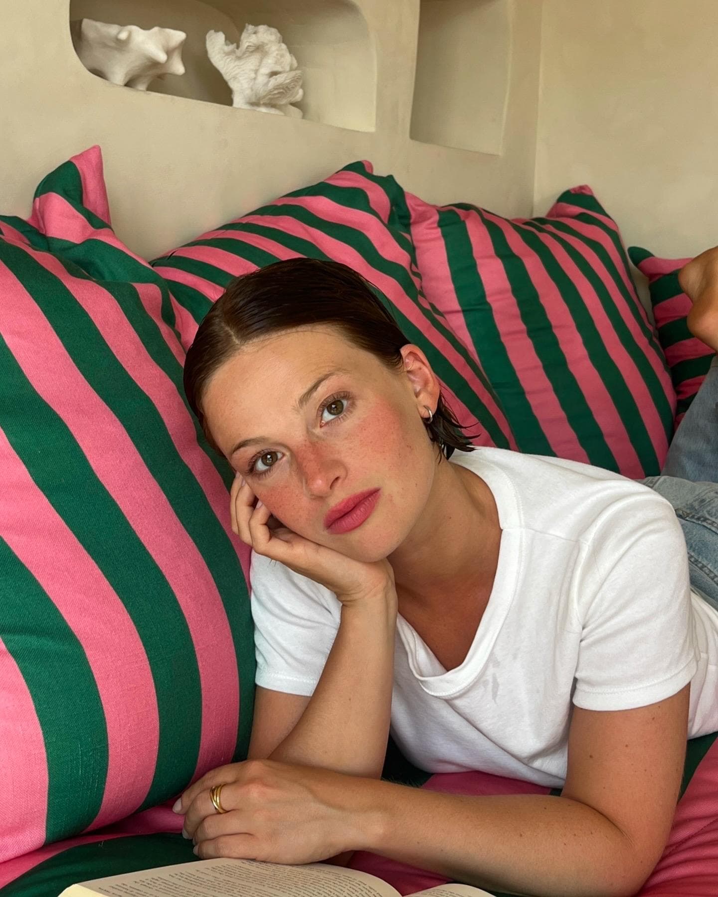 Une femme aux cheveux bruns courts est allongée sur des oreillers rayés roses et verts, posant sa tête sur sa main et regardant la caméra. Elle porte un t-shirt blanc et se trouve à côté d'un livre ouvert. Des étagères et un décor se trouvent à l'arrière-plan.