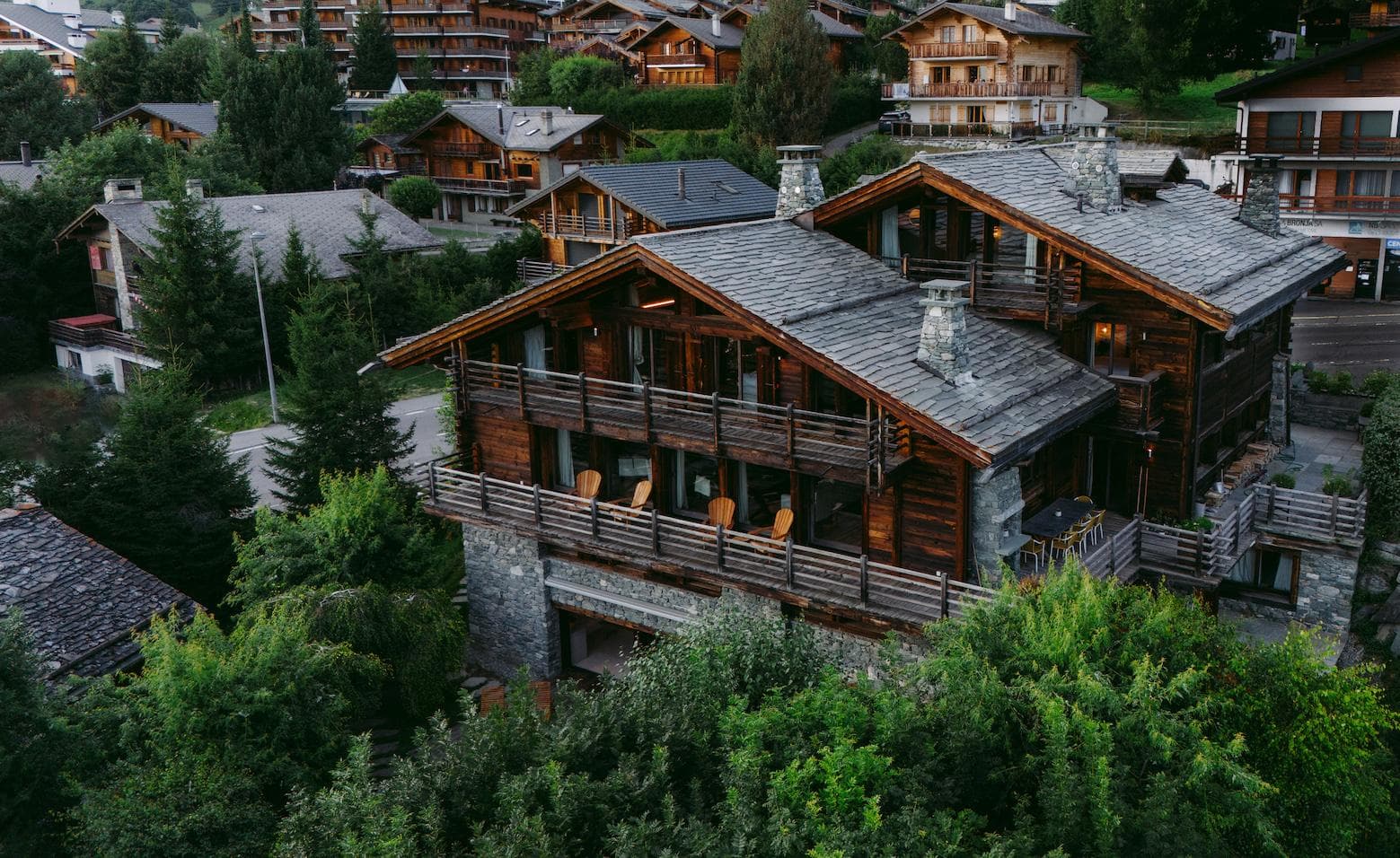 A large, rustic wooden chalet with stone accents is nestled among green trees, surrounded by similar alpine-style houses in a mountainous village setting.