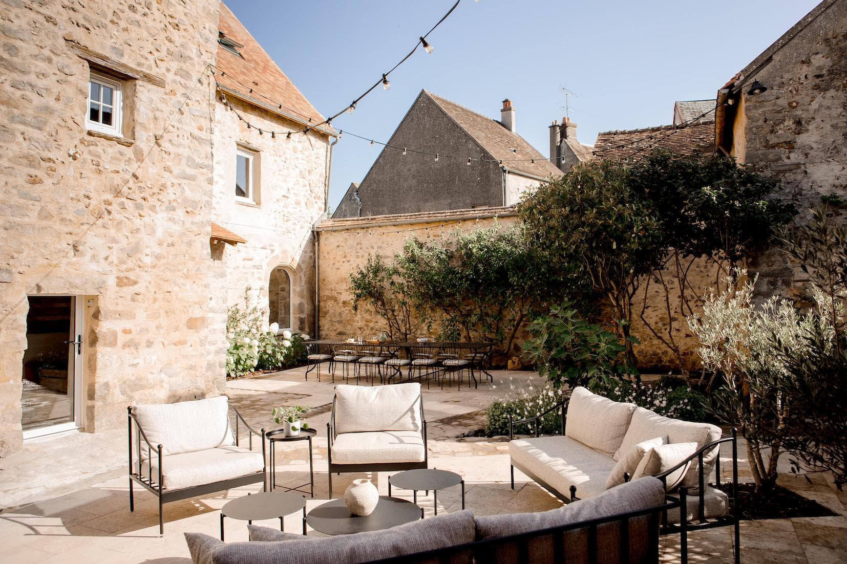 Une cour en pierre éclairée par le soleil, avec des canapés d'extérieur de couleur crème, des tables rondes noires et des guirlandes lumineuses au-dessus de la tête. Des plantes vertes tapissent les murs et de vieux bâtiments en pierre entourent le coin salon.