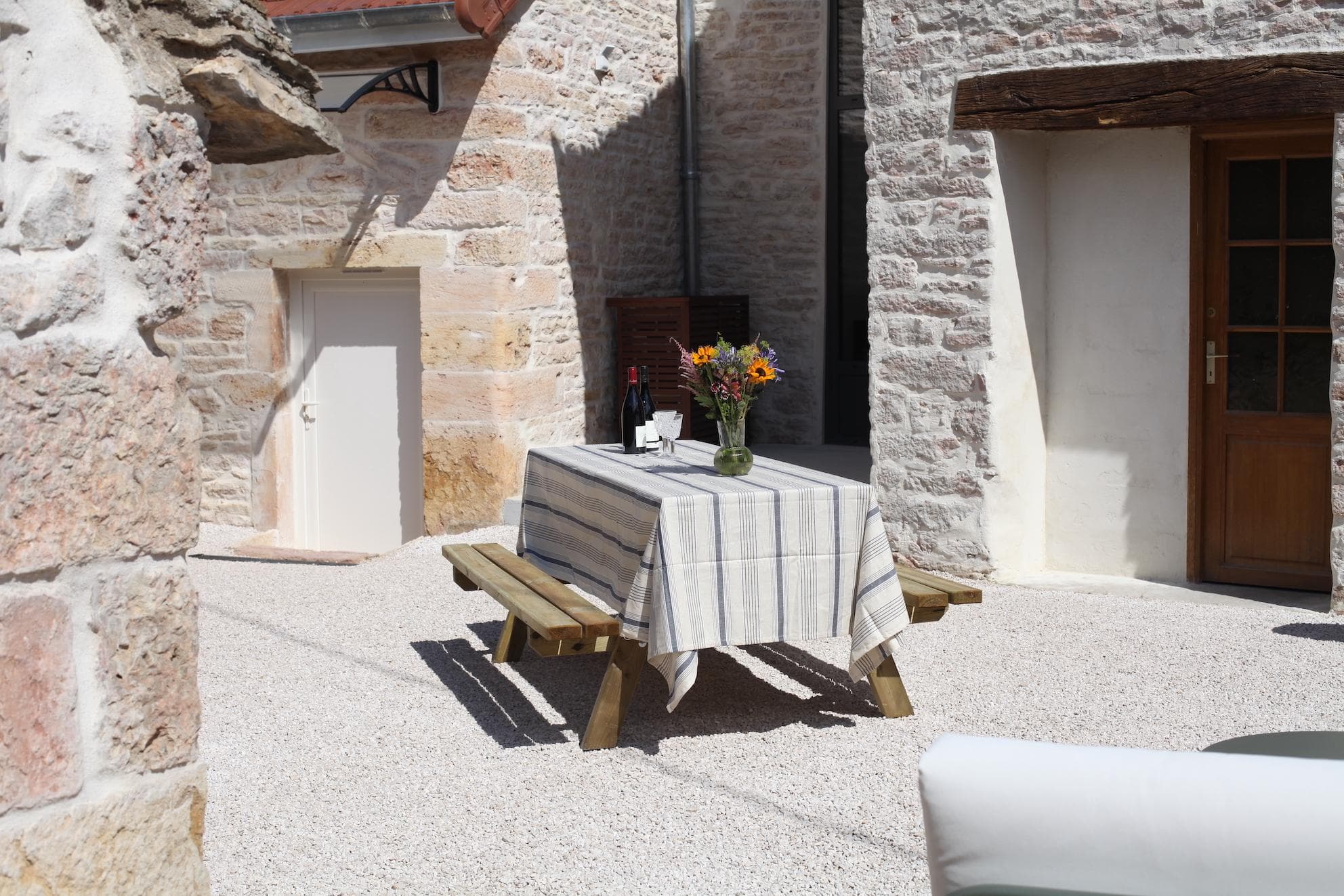 A wooden picnic table with a striped tablecloth, flowers, and wine sits in a sunlit stone courtyard near a wooden door and small white door.