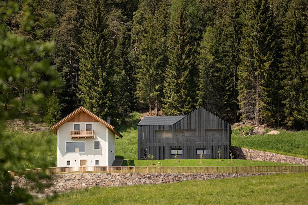 Deux maisons modernes, l'une blanche avec un toit en bois et un balcon, l'autre gris foncé avec un design épuré, sont assises côte à côte sur l'herbe verte devant une forêt dense de grands pins.
