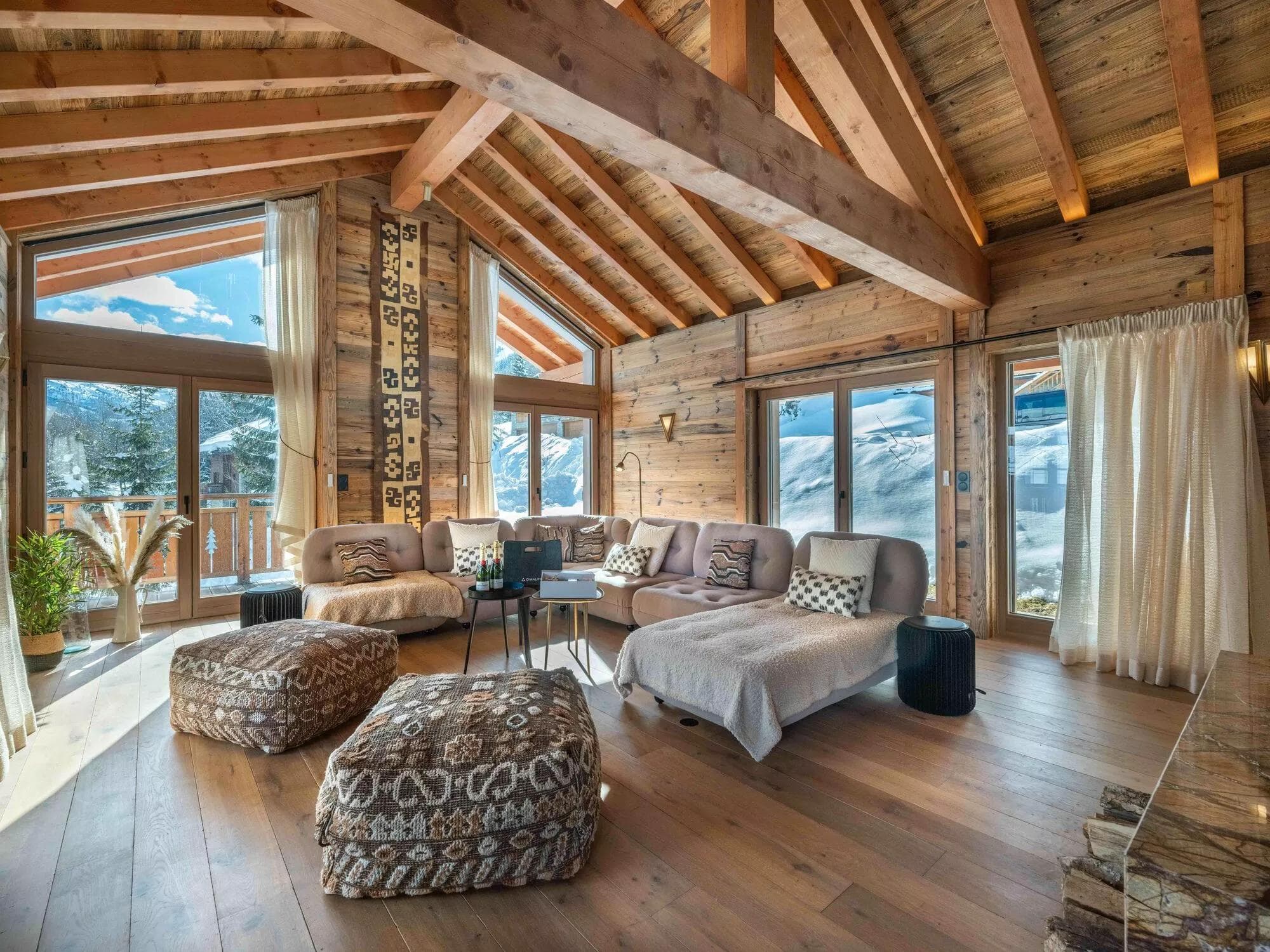 Salon spacieux d'une cabane de montagne avec de grandes fenêtres, des poutres en bois, un canapé sectionnel avec des coussins à motifs et des ottomans, des plantes et une vue sur les sommets enneigés à l'extérieur. La lumière naturelle remplit cet espace rustique et confortable.