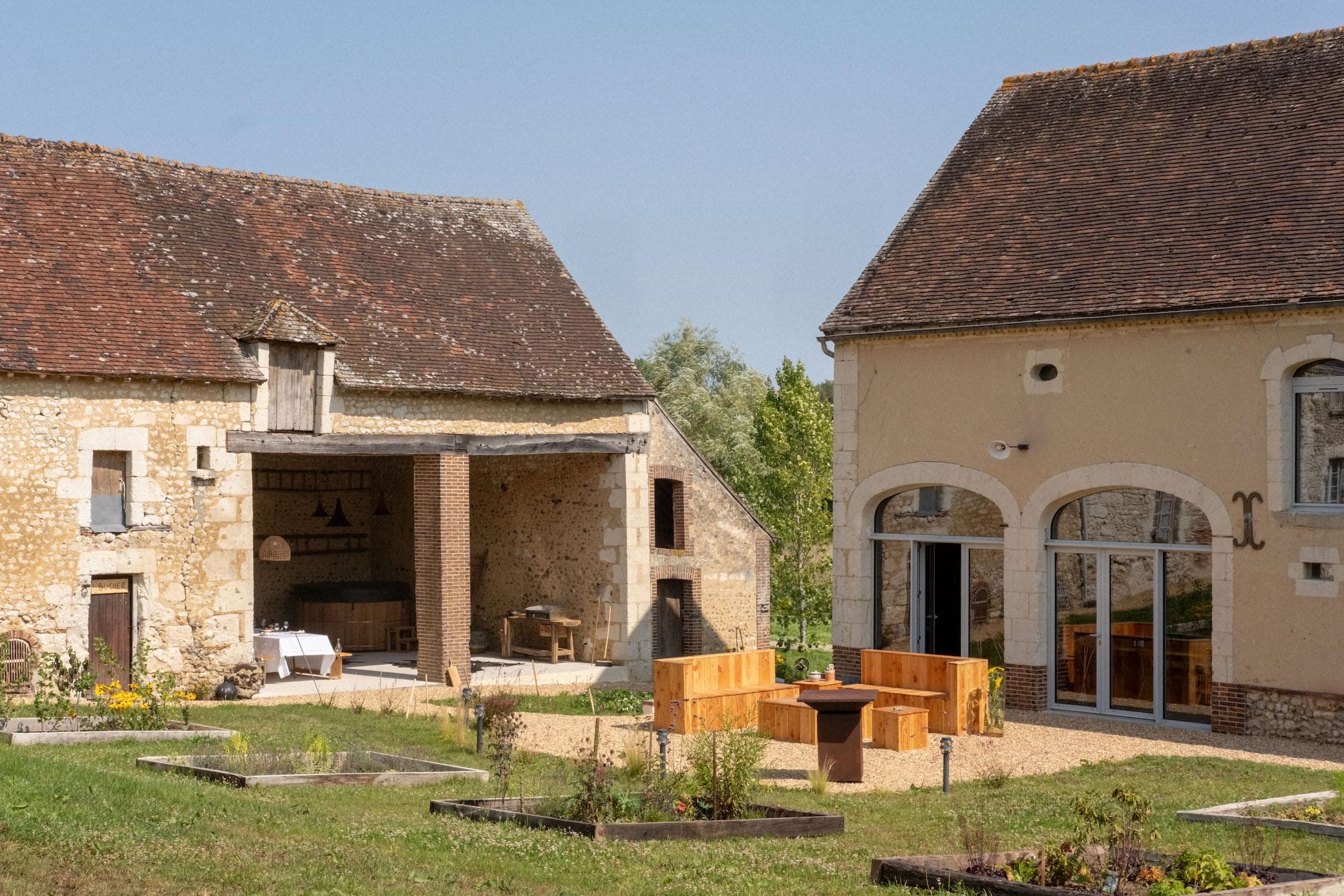A rustic courtyard with two stone buildings featuring wooden and brick details, a patio area with wooden benches and a fire pit, surrounded by grassy garden beds and flowers under a clear blue sky.