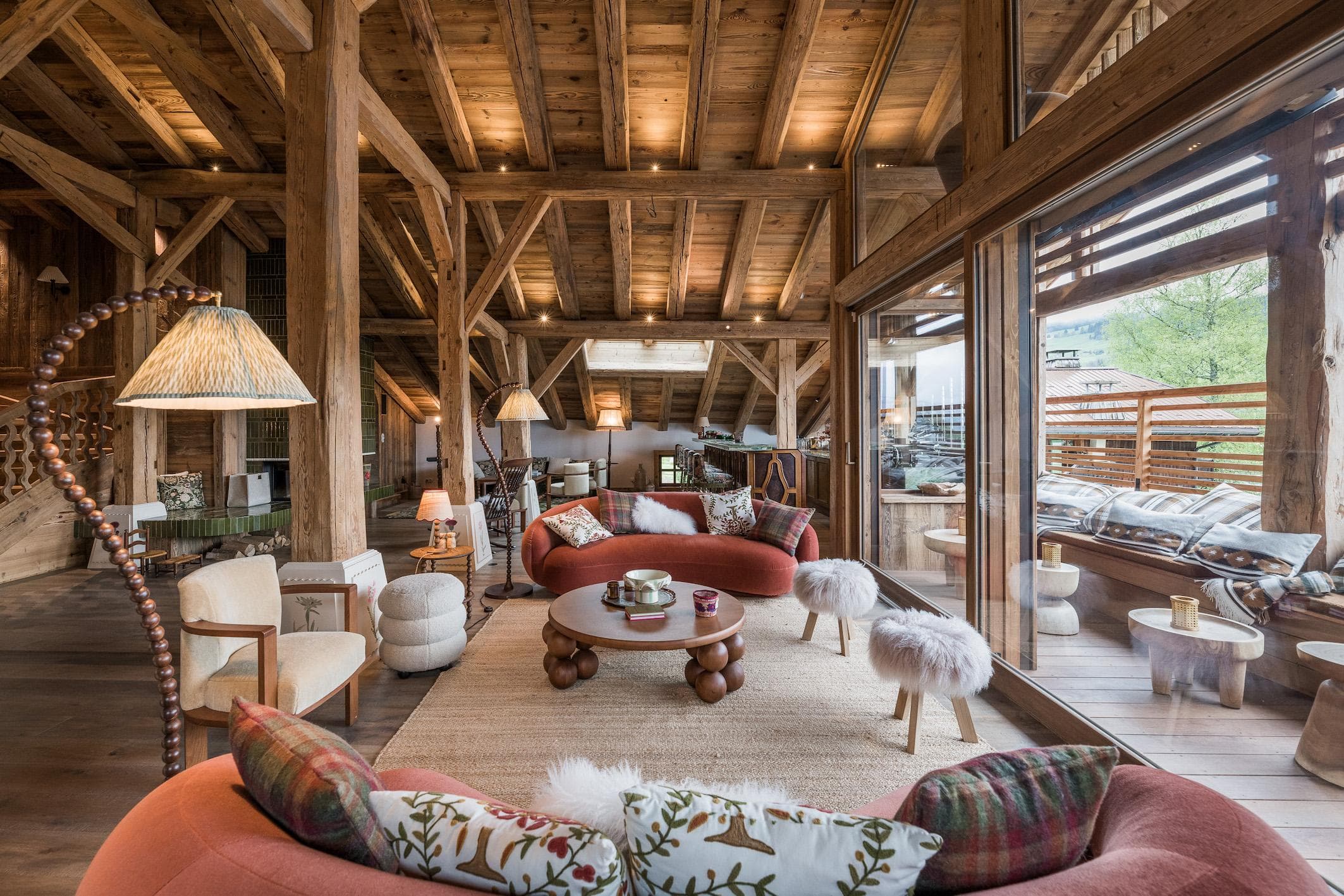 Un salon rustique et confortable avec des poutres en bois apparentes, un éclairage chaleureux, deux canapés rouges incurvés, des tabourets rembourrés, une table basse ronde et de grandes portes vitrées qui s'ouvrent sur une terrasse en bois avec des sièges d'extérieur.