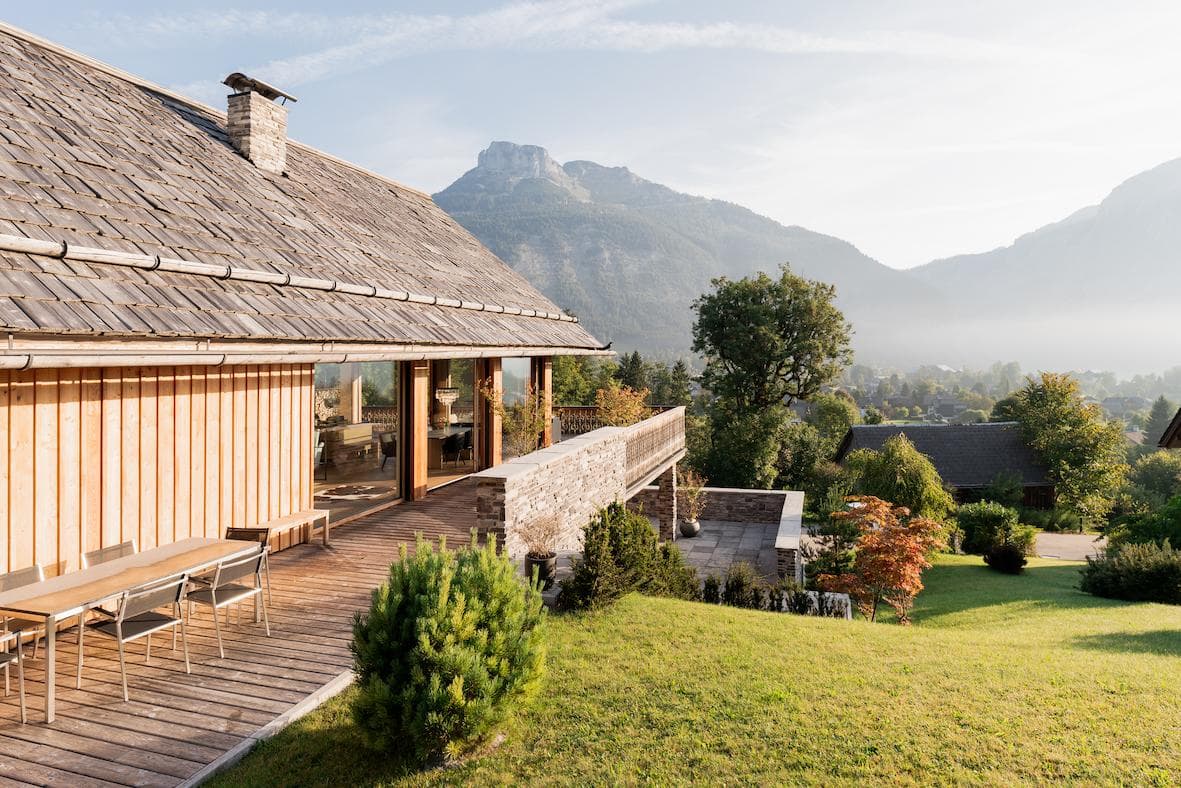 A modern wooden house with a large deck overlooks a lush green lawn and a scenic mountain landscape under a clear sky, with trees and distant houses visible in the background.