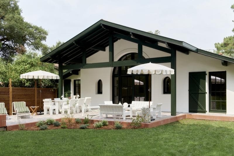 A modern white house with dark green trim features a covered patio with white outdoor furniture and two white umbrellas, surrounded by a green lawn and landscaped garden beds.
