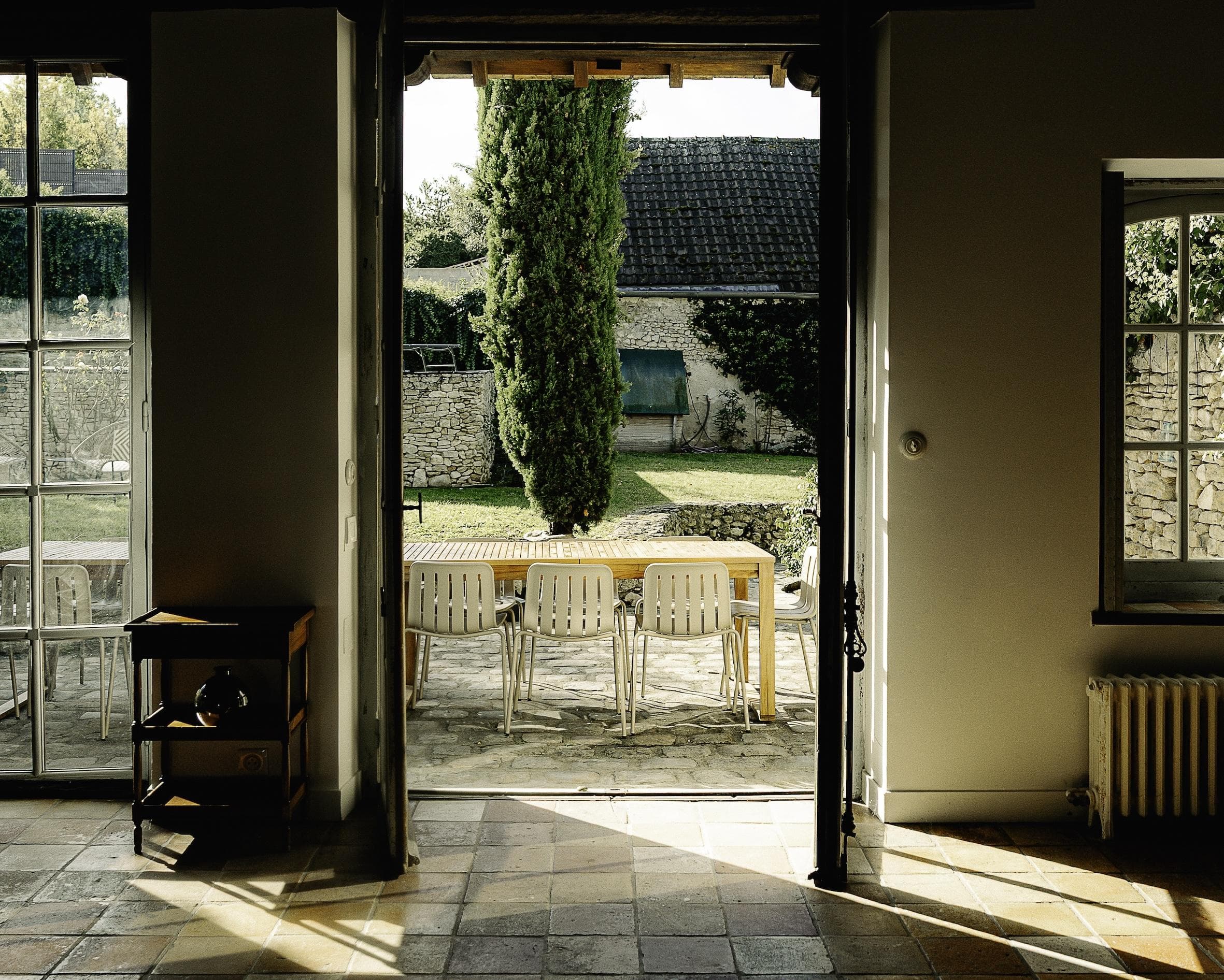 Sunlit view through open double doors to a patio with a wooden table and white chairs, overlooking a garden with green grass, a tall tree, and stone walls in the background.