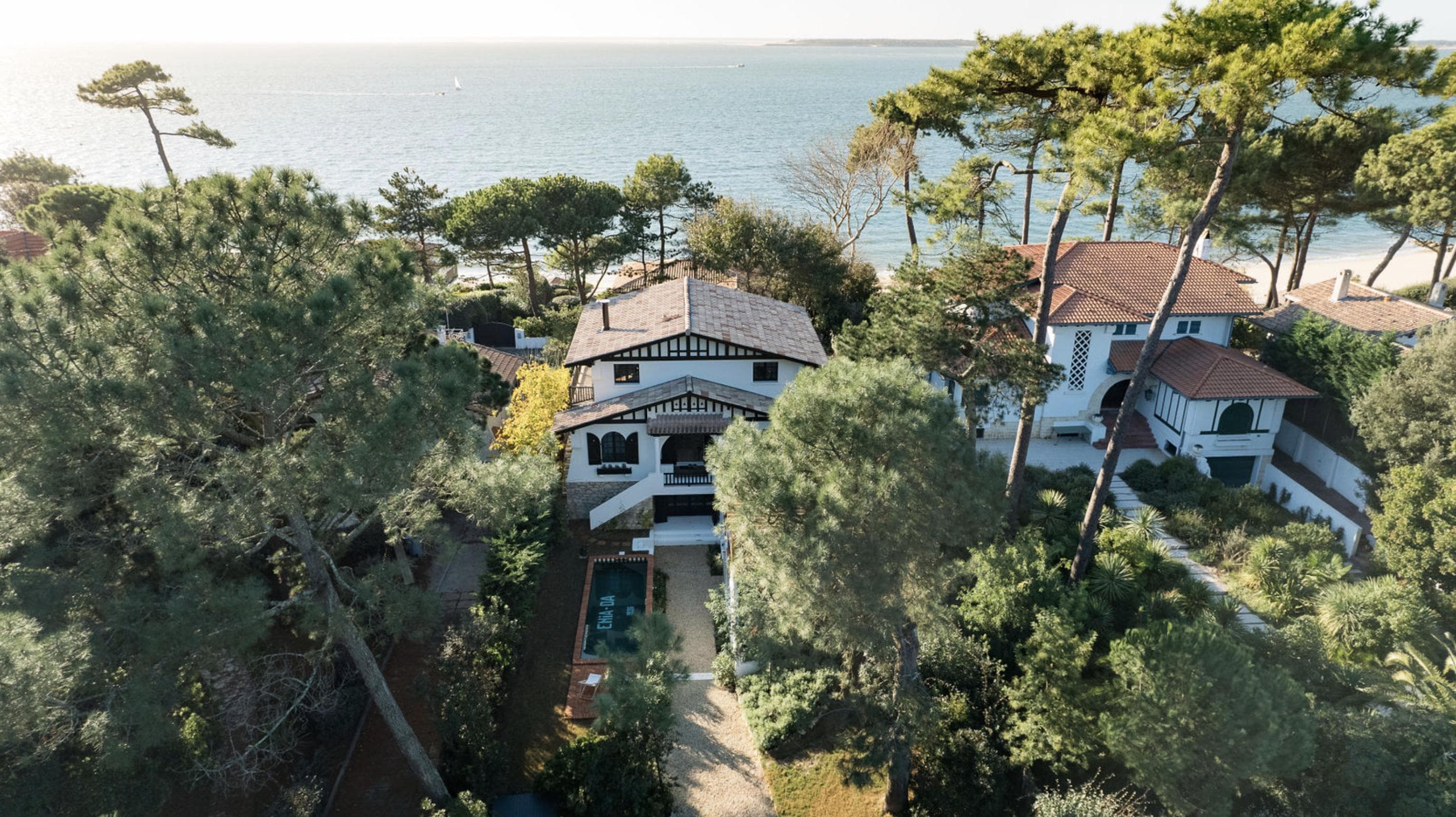 Aerial view of a coastal house surrounded by tall trees, with a swimming pool in the backyard and the ocean visible in the background. Neighboring houses are visible on either side.