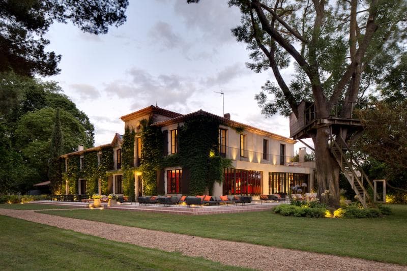 A large two-story house with ivy-covered walls, large windows, and warm outdoor lighting sits in a landscaped garden with a gravel path. A wooden treehouse is built around a tree on the right side of the image.