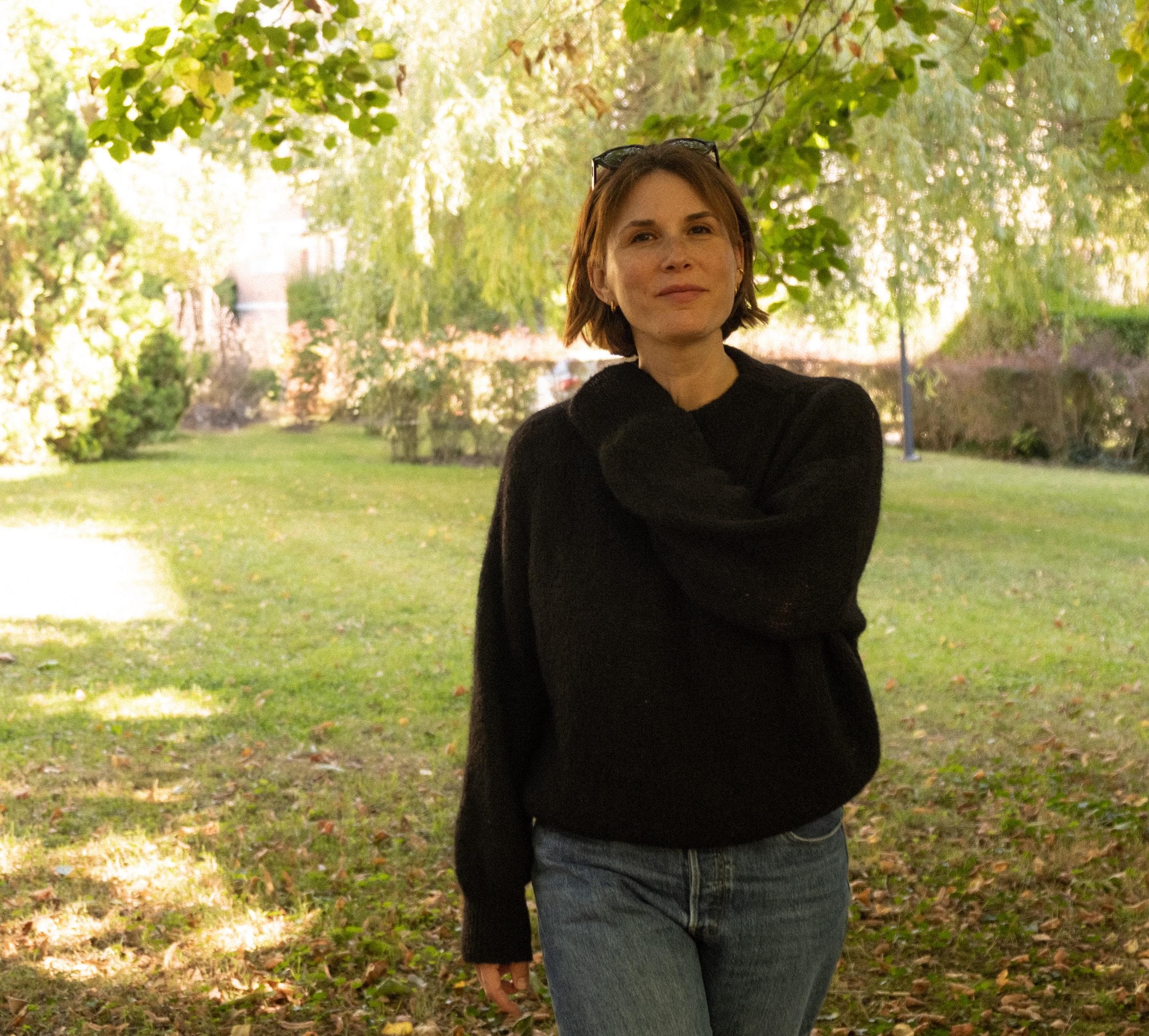 Une personne aux cheveux bruns courts, vêtue d'un pull sombre et d'un jean bleu, se tient à l'extérieur, dans un parc herbeux et ensoleillé, entouré d'arbres et de verdure.