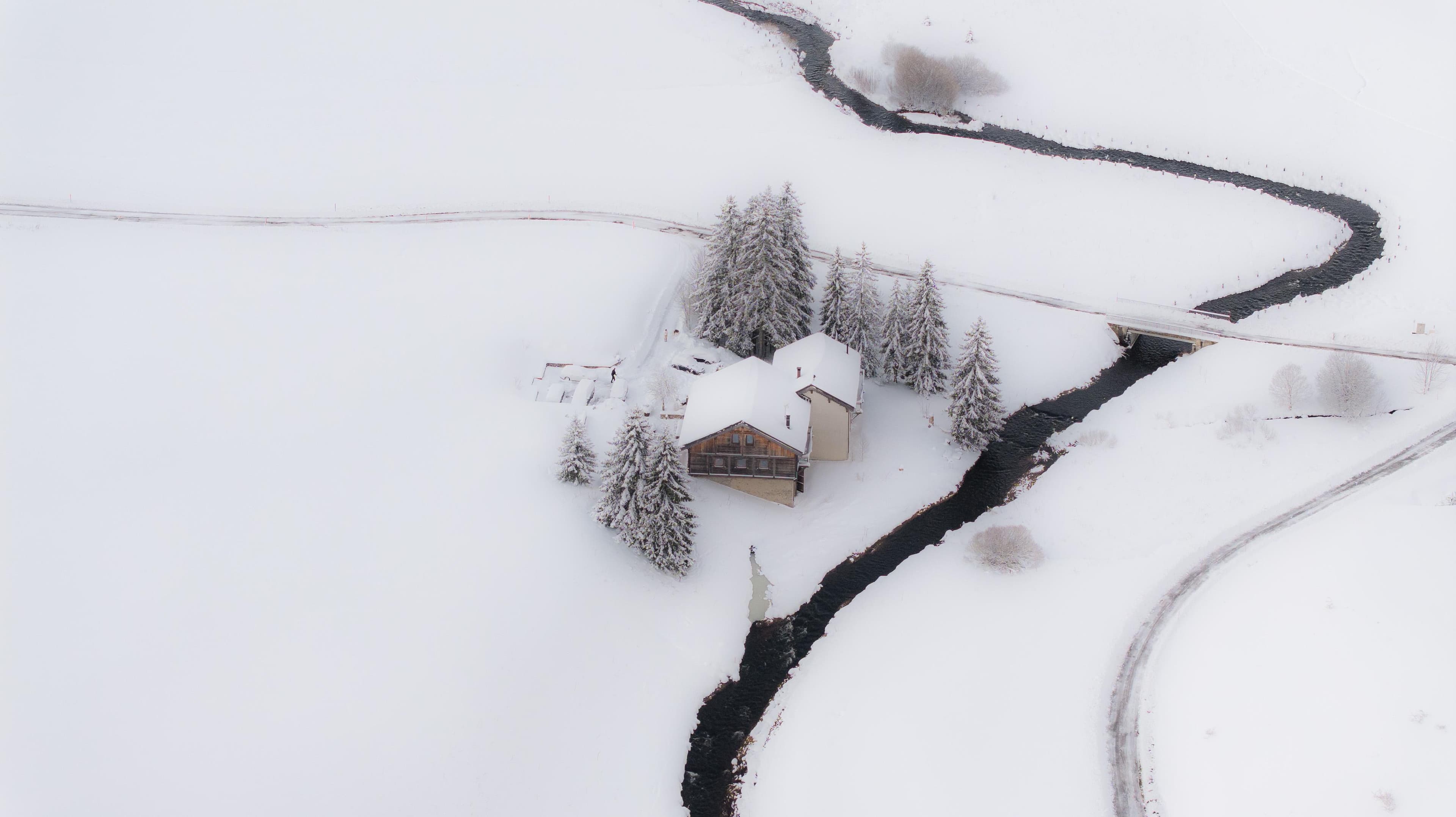 A small house surrounded by snow-covered trees sits beside a narrow, winding river in a snowy landscape, with a bridge crossing the river nearby.