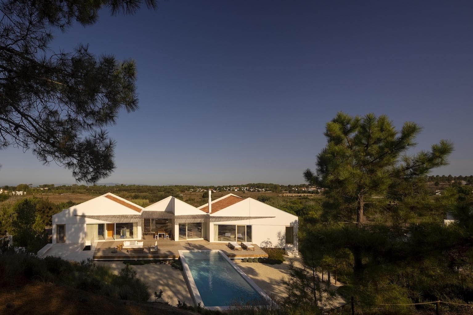 A modern white villa with large windows and a central swimming pool, surrounded by trees and greenery, under a clear blue sky at sunset.