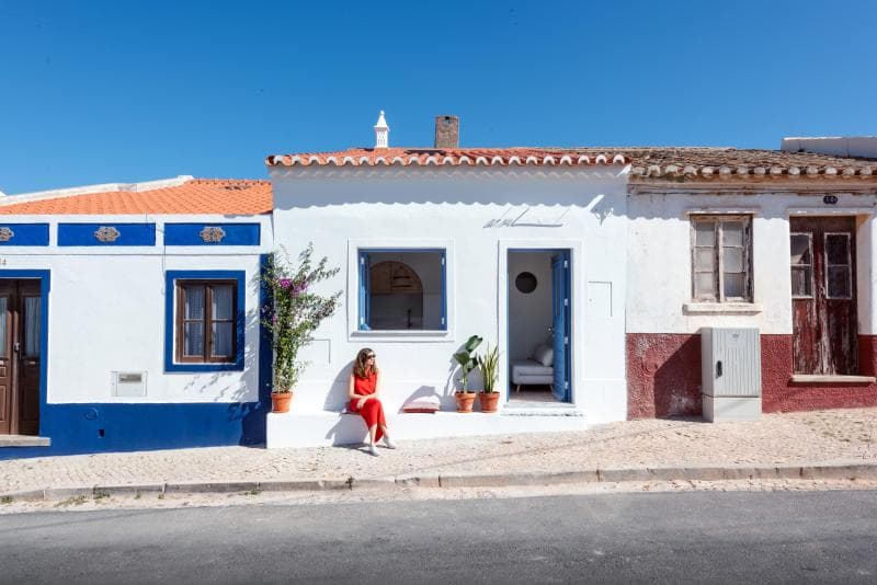 Une femme vêtue d'une robe rouge est assise sur un banc blanc à l'extérieur d'une rangée de maisons méditerranéennes traditionnelles et colorées, sous un ciel bleu clair. Les bâtiments ont des accents bleus, blancs et rouges vifs.
