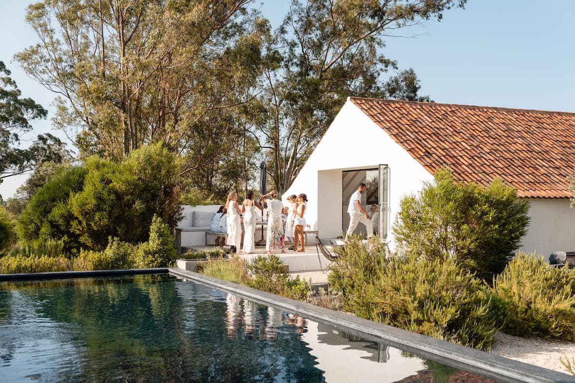 A group of people dressed in white gather on a patio outside a modern house with a red-tiled roof, surrounded by greenery, near a reflective swimming pool on a sunny day.
