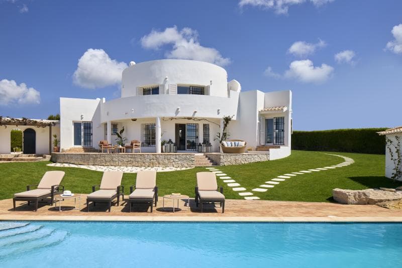 Une villa blanche moderne à l'architecture arrondie, avec de grandes fenêtres et un patio spacieux donnant sur une piscine avec quatre chaises longues lors d'une journée ensoleillée. La pelouse environnante est soigneusement aménagée avec des marches d'escalier.