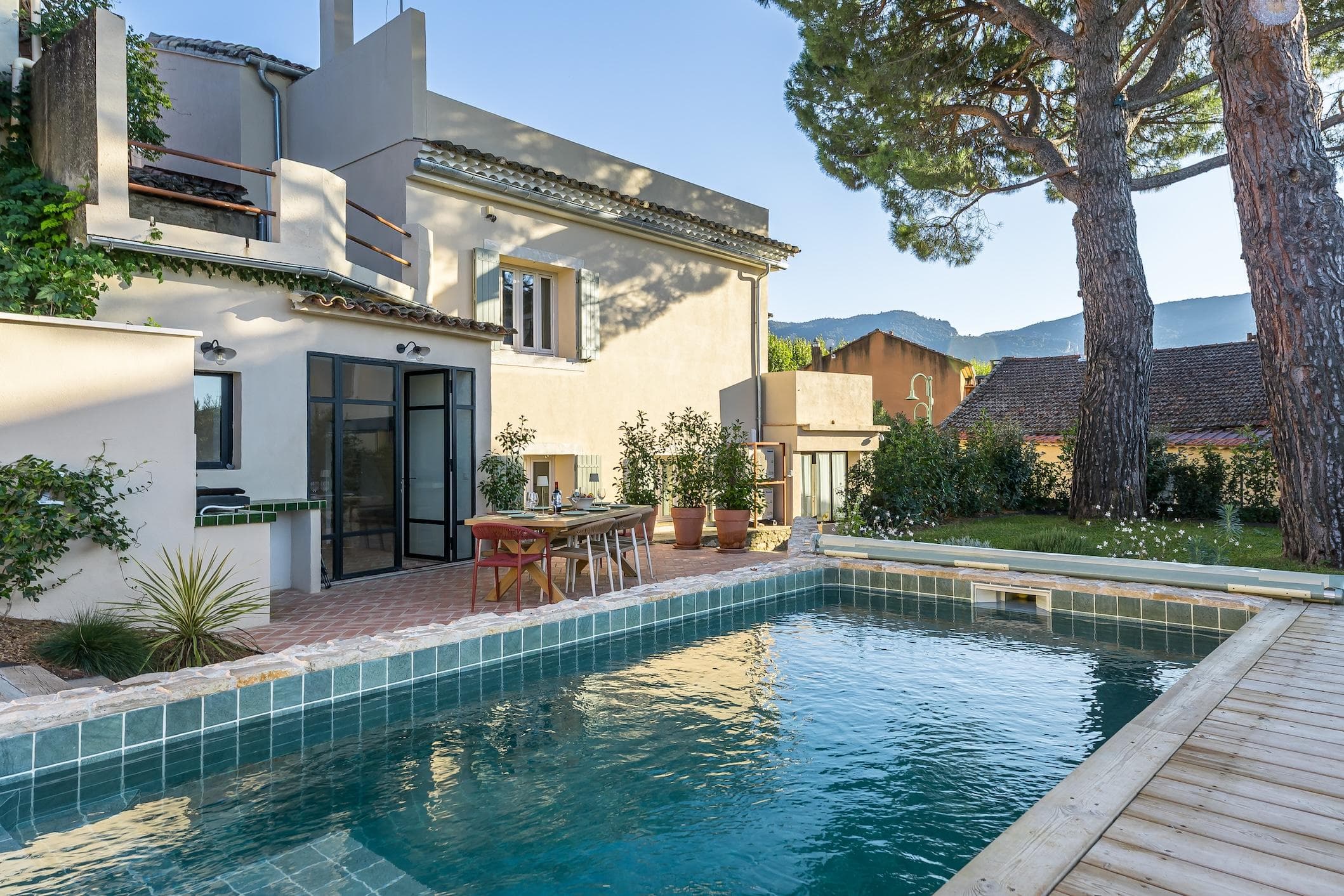 Maison moderne avec de grandes fenêtres et un balcon, près d'une piscine rectangulaire et d'une table à manger extérieure, entourée d'arbres, de plantes et de montagnes en arrière-plan lors d'une journée ensoleillée.