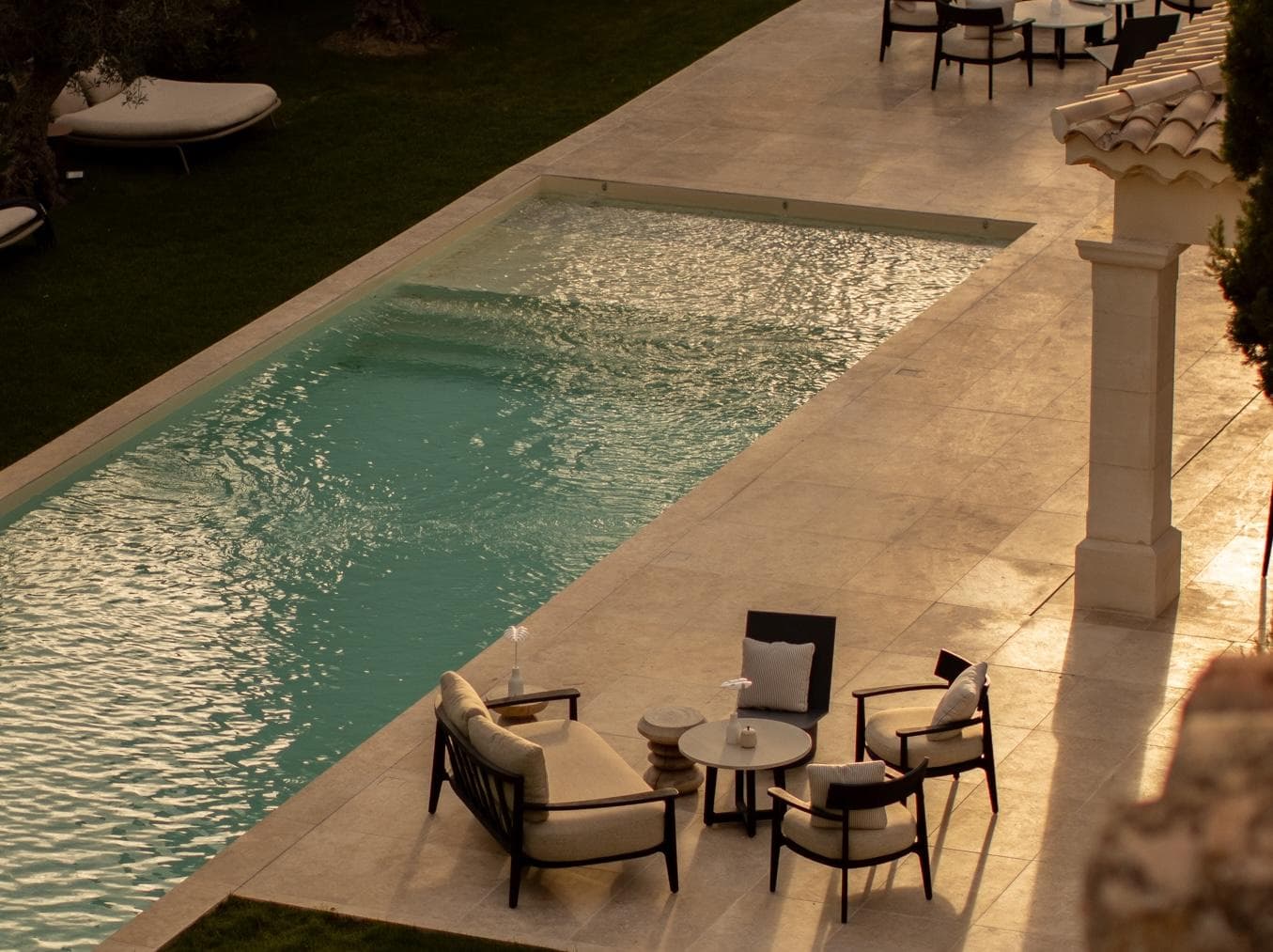 Outdoor seating area with cushioned chairs and small tables beside a rectangular swimming pool, surrounded by beige tiles and soft sunlight. Some tables have drinks on them. The scene looks calm and inviting.