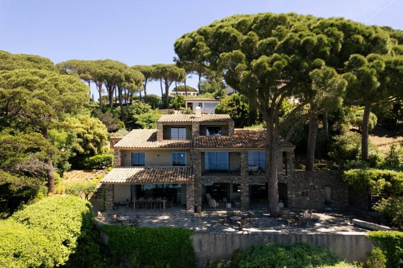 Une grande maison en pierre avec un toit en tuiles se trouve au milieu de grands pins et de verdure, avec un patio spacieux et un espace pour s'asseoir à l'extérieur, sous un ciel bleu clair.