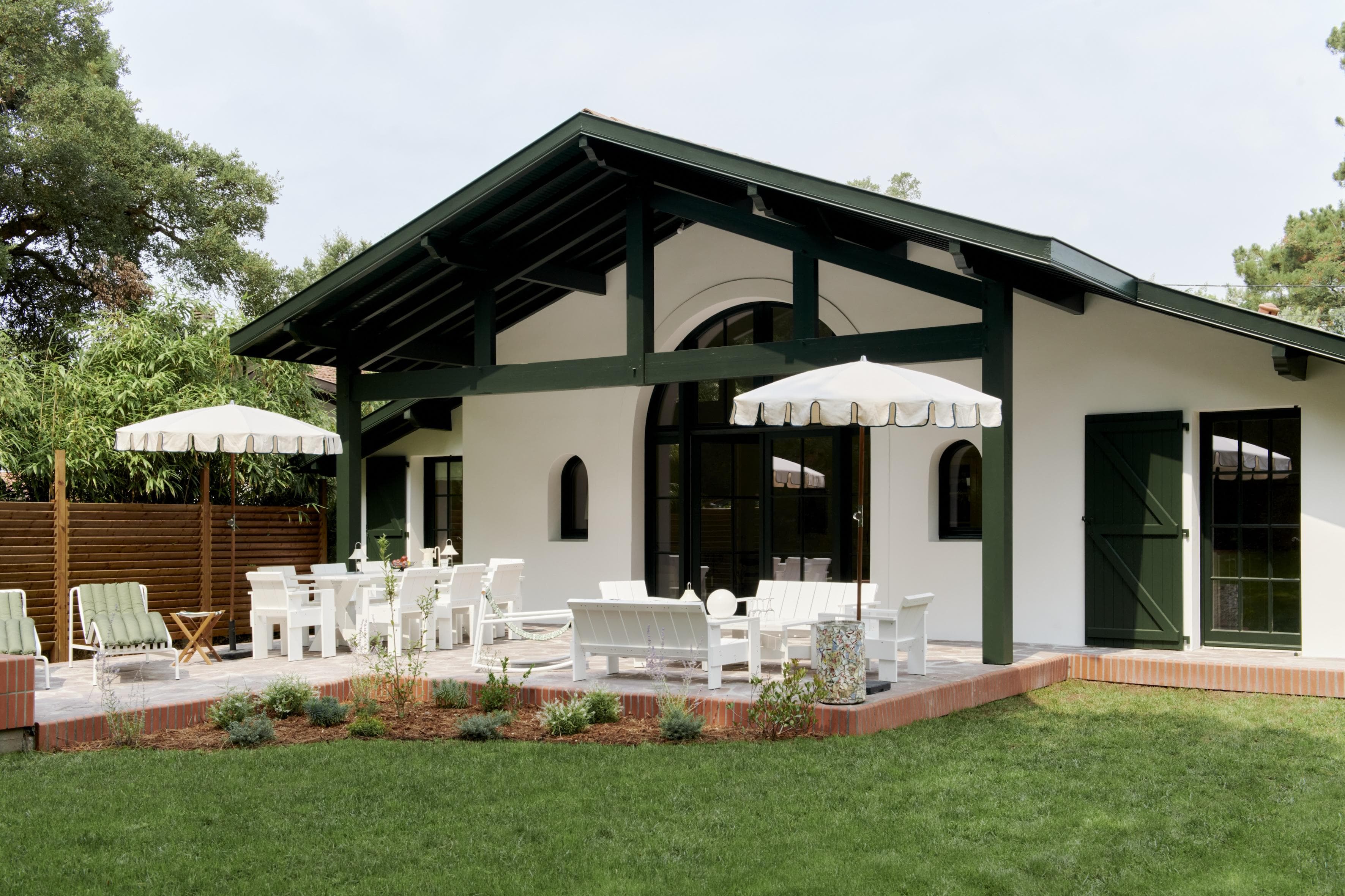 Une maison blanche avec des bordures vert foncé dispose d'un patio couvert, de meubles d'extérieur blancs, de deux parasols blancs et d'un jardin paysager, le tout situé dans une cour herbeuse avec des arbres et une clôture en bois.