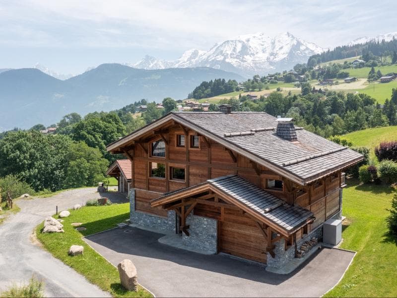 A large wooden chalet with stone accents sits on a green hillside, surrounded by trees and mountains, with snow-capped peaks visible in the background under a partly cloudy sky.