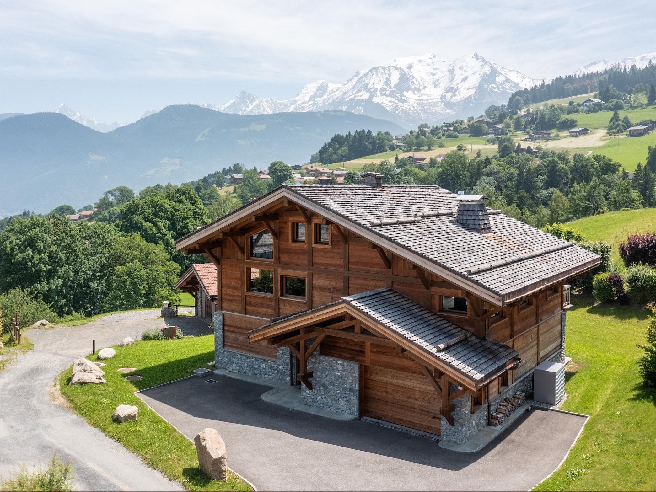 Un grand chalet en bois avec des accents de pierre se trouve sur une colline verdoyante, entouré d'arbres et de montagnes, avec des sommets enneigés visibles en arrière-plan sous un ciel partiellement nuageux.