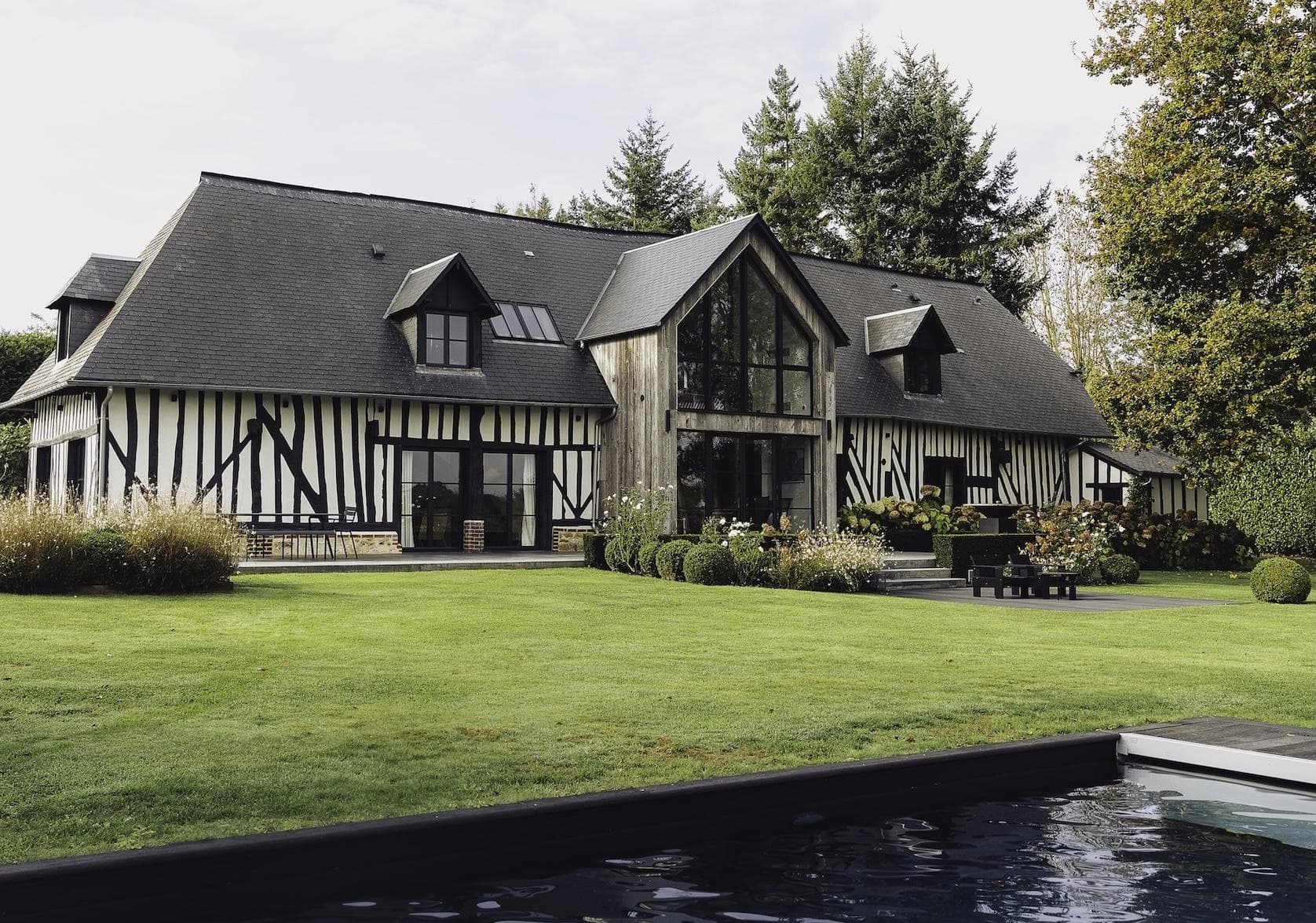 A large, modern Tudor-style house with black timber framing and white walls sits behind a neatly manicured lawn, garden, and a swimming pool, surrounded by tall trees.