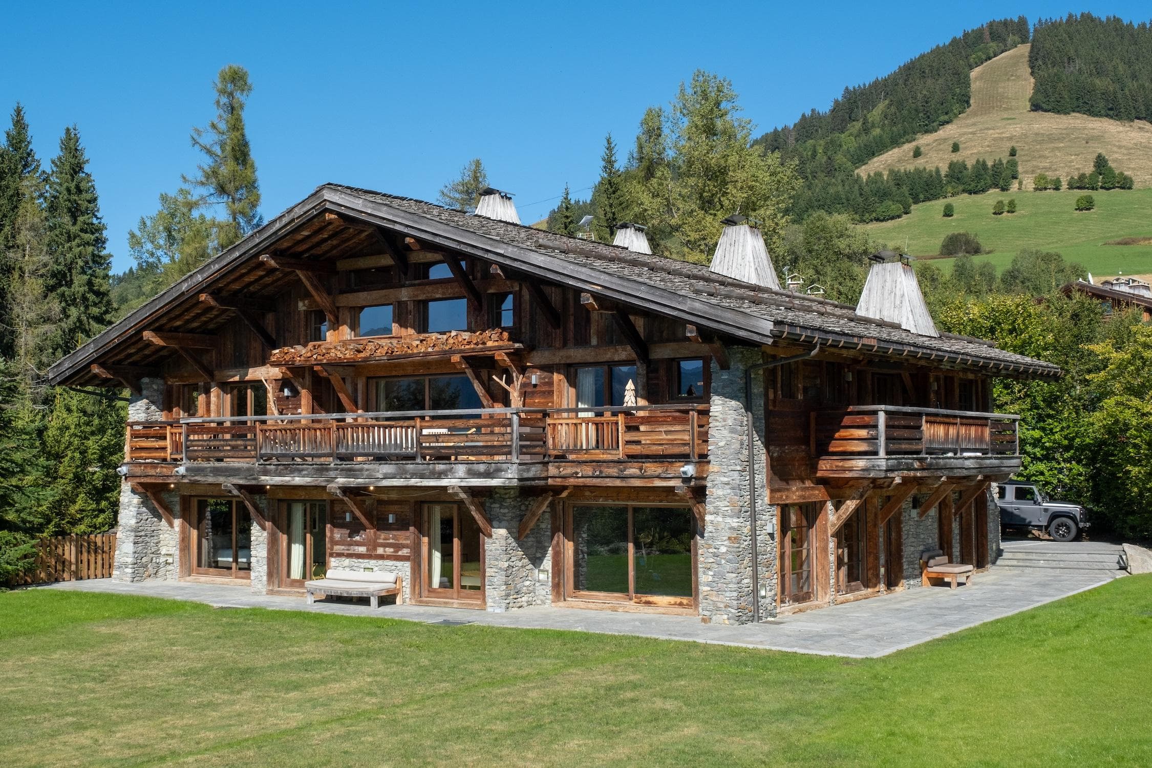 A large wooden chalet with stone accents and balconies stands on a grassy lawn, surrounded by trees, with green hills and a clear blue sky in the background.