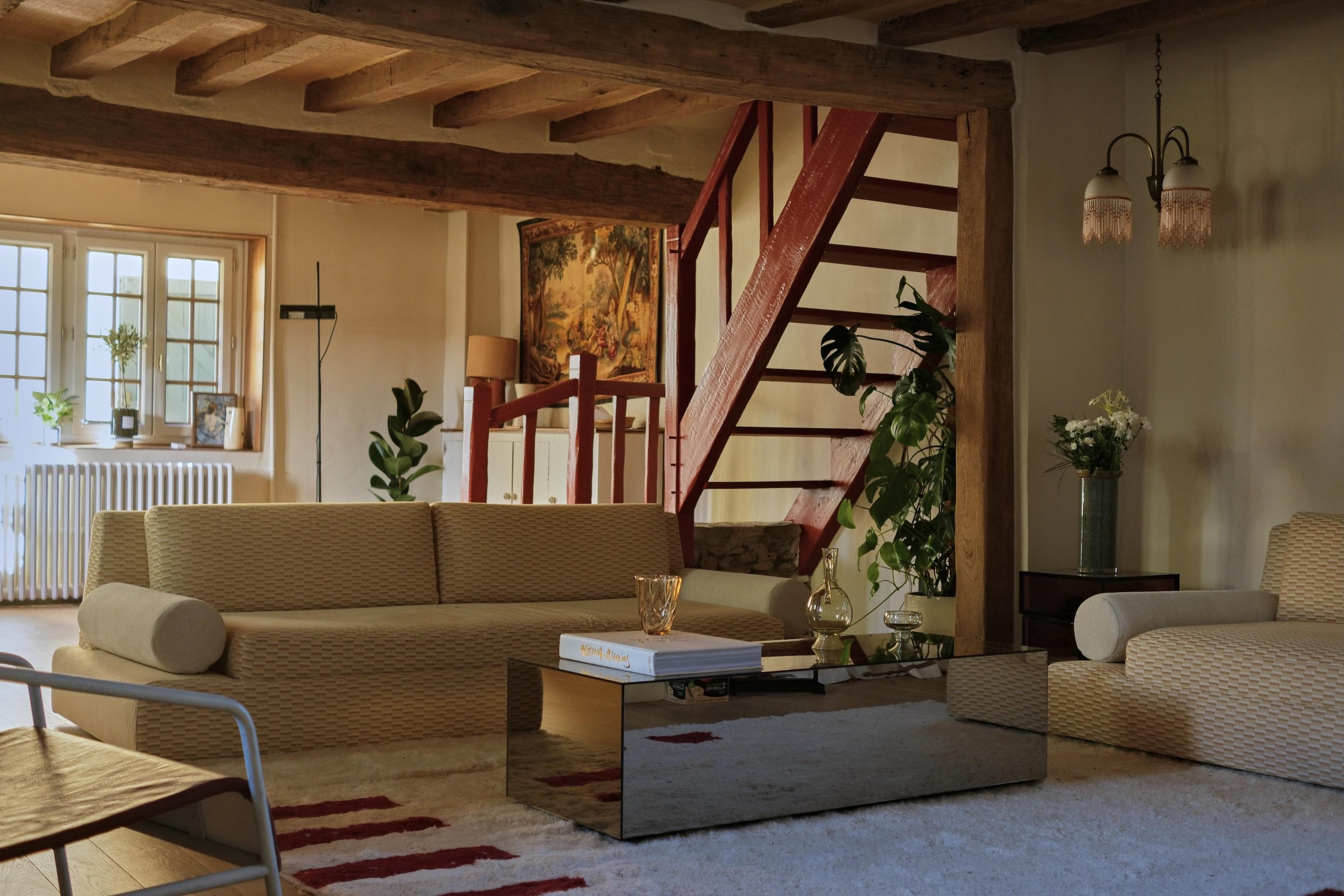 Un salon confortable avec des canapés beiges, une table basse en verre, des plantes d'intérieur, un plafond à poutres apparentes et un escalier rouge menant à l'étage. La lumière chaude pénètre par de grandes fenêtres, créant une atmosphère accueillante.