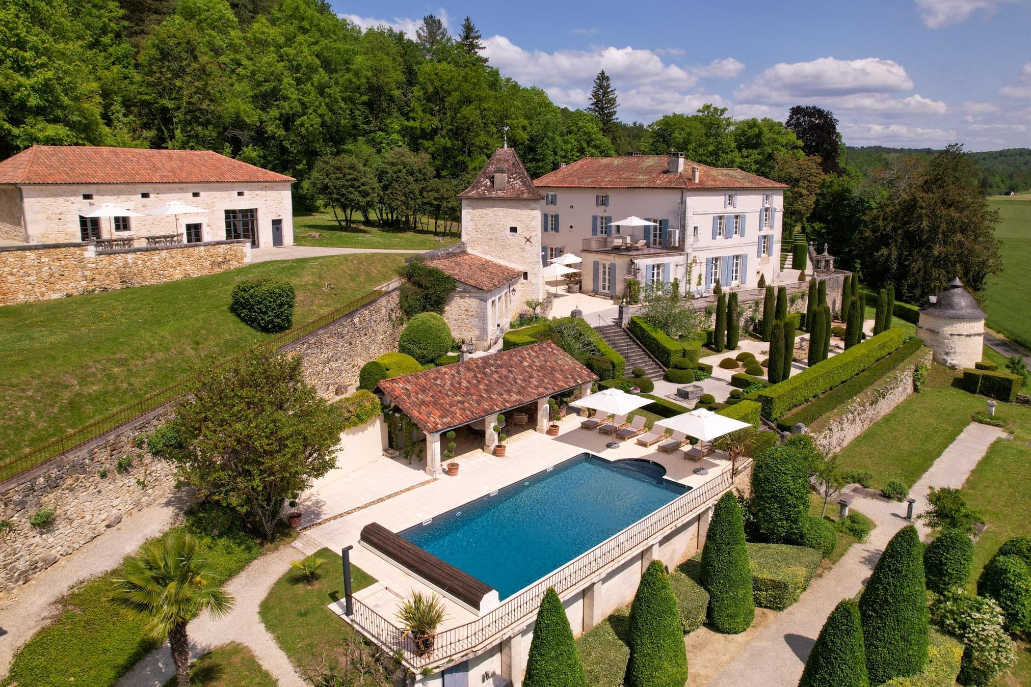 A large, elegant estate with a swimming pool, manicured gardens, stone buildings, and lush greenery, set on a hillside under a partly cloudy blue sky.