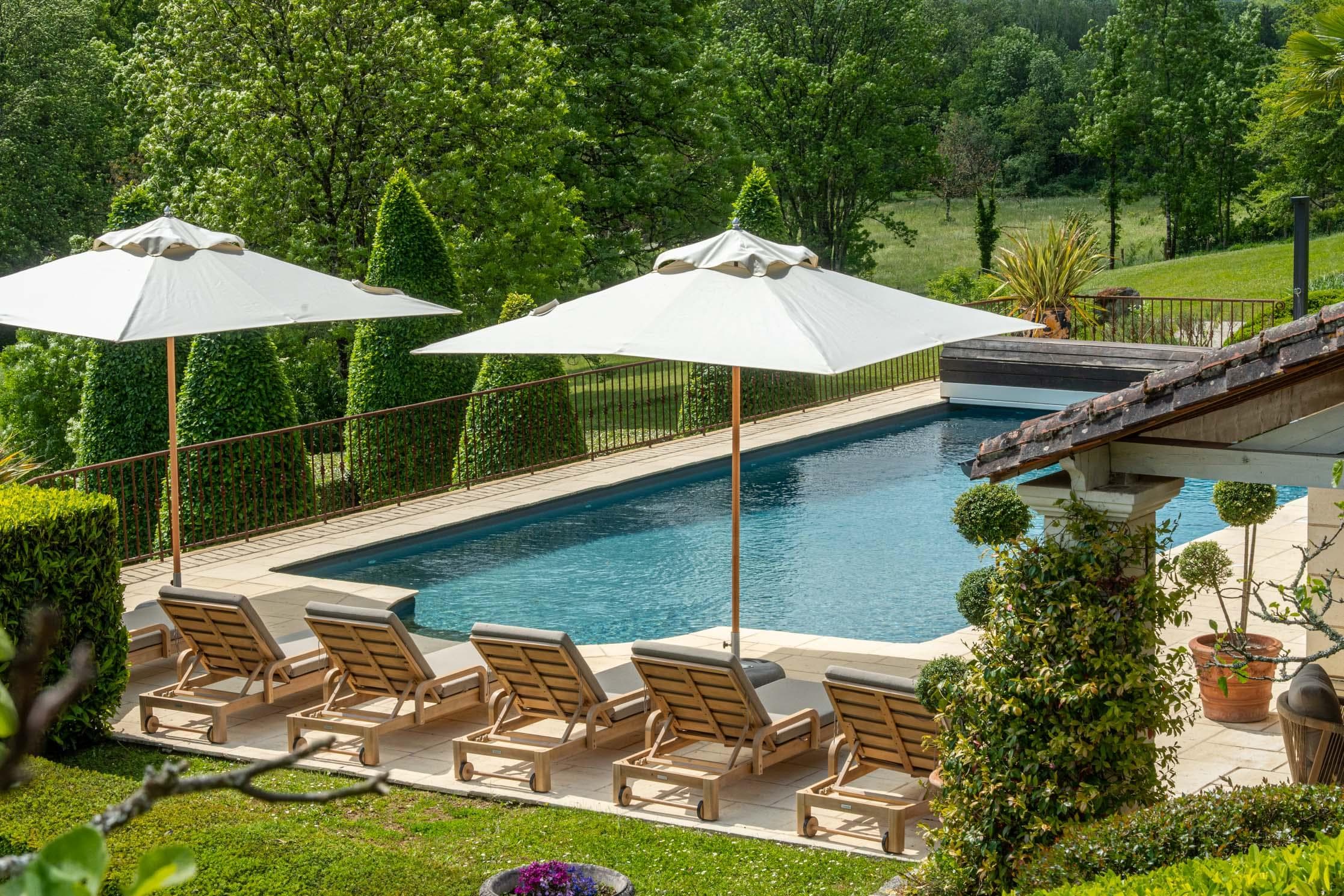 A rectangular outdoor swimming pool with six wooden lounge chairs and two large white umbrellas, surrounded by greenery and trees in a landscaped garden.