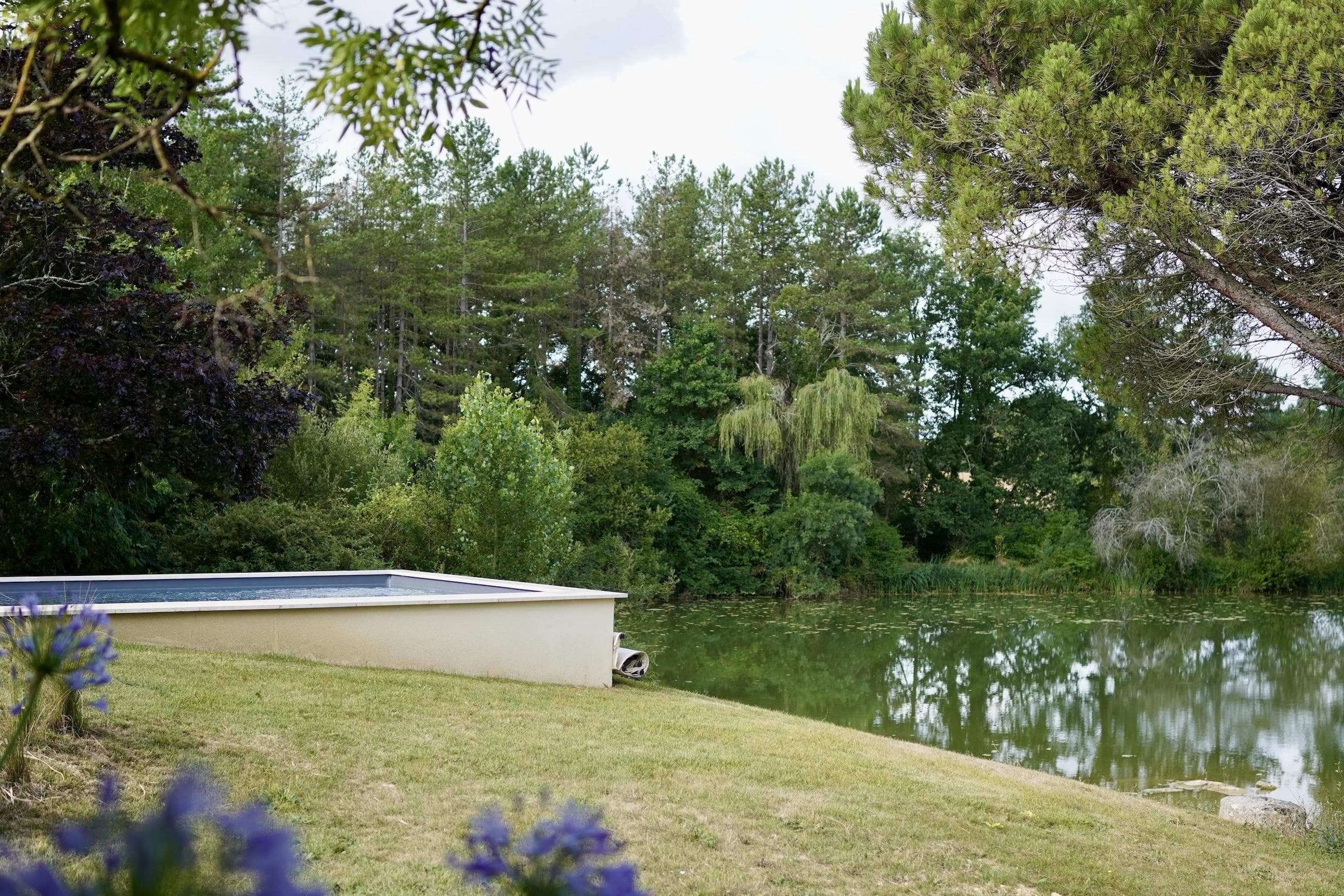A small rectangular pool sits on a grassy slope beside a calm, tree-lined lake, surrounded by lush greenery and flowering plants under a cloudy sky.