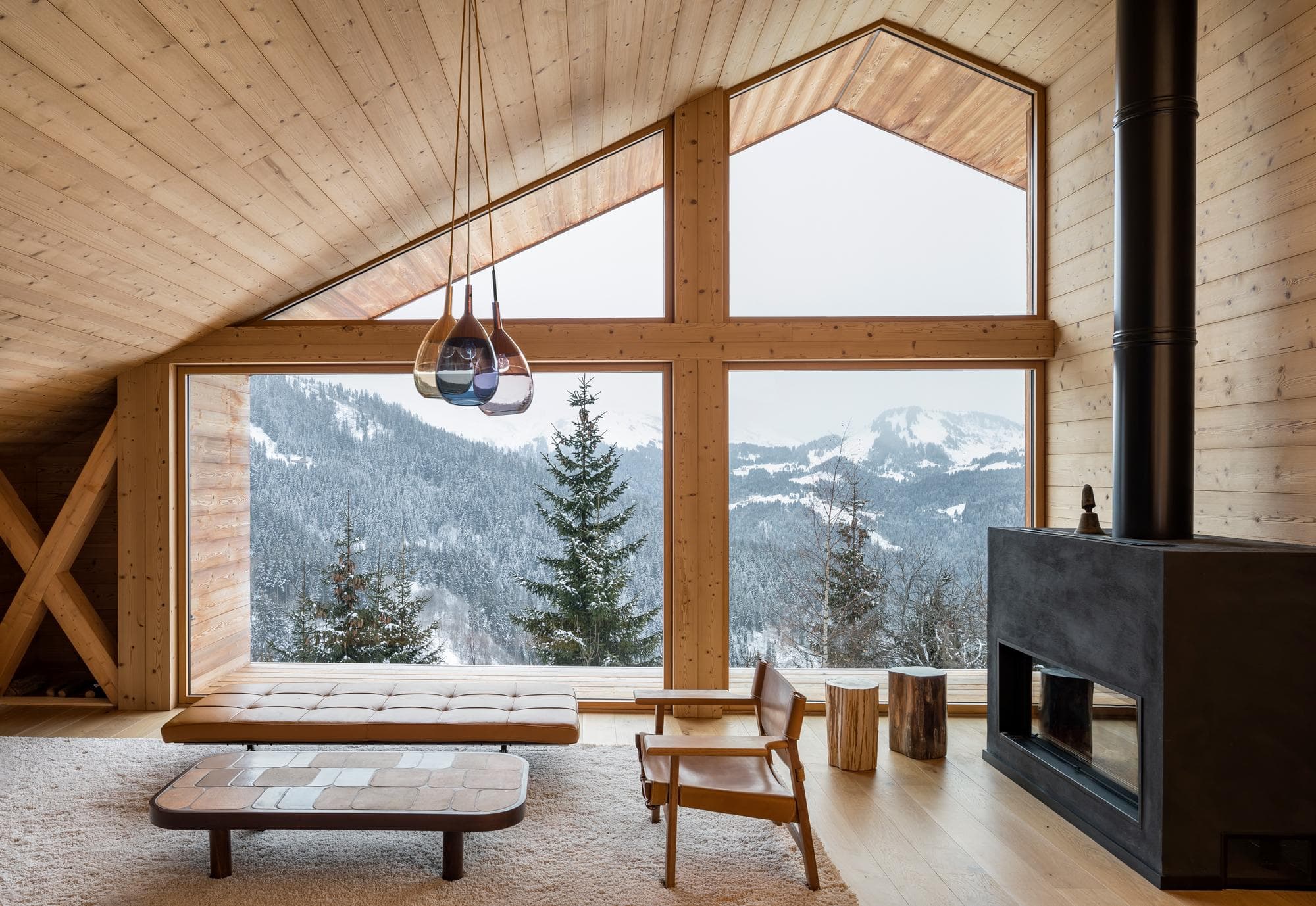 A modern wooden cabin interior with large windows overlooking a snowy mountain landscape, featuring a cozy seating area, minimalist furniture, a black fireplace, and natural wood finishes.